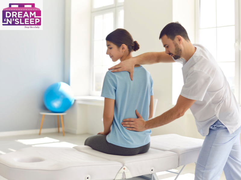 Woman sitting on bed with back pain while man helps adjust her back for support